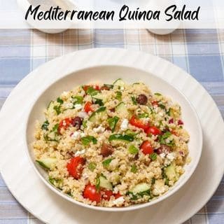 A bowl of quinoa salad with tomatoes, cucumbers, olives, and feta on a checkered tablecloth, accompanied by salt, pepper shakers, and a jug of dressing.