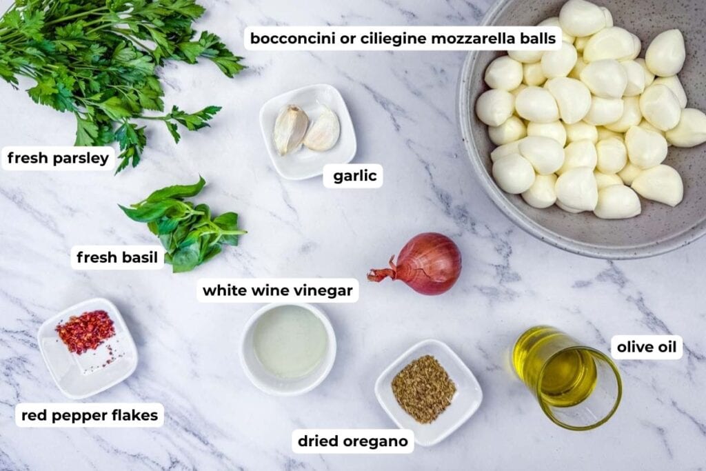 Ingredients for a mozzarella salad are arranged on a marble surface, including mozzarella balls, parsley, basil, garlic, shallot, vinegar, olive oil, oregano, and red pepper flakes.