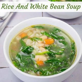 A bowl of rice and white bean soup with spinach and diced carrots, placed on a light-colored surface.