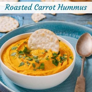 Bowl of roasted carrot hummus garnished with herbs and topped with a round cracker, served on a blue plate with a spoon and additional crackers in the background.