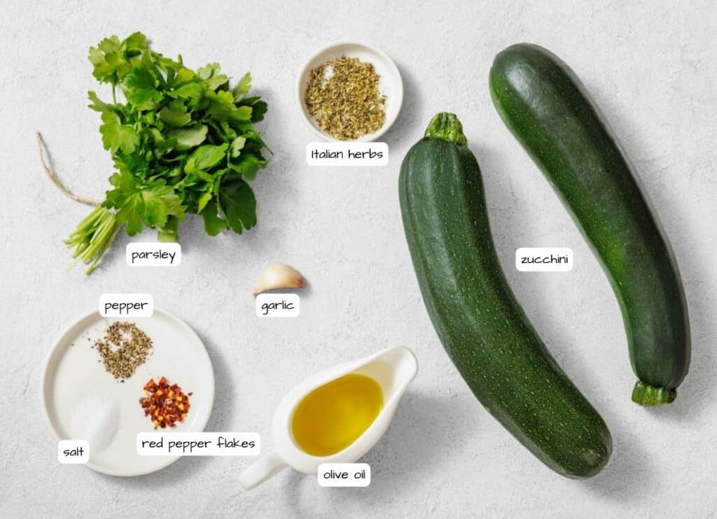 Ingredients for a recipe arranged on a white surface: parsley, Italian herbs, garlic, two zucchinis, pepper, salt, red pepper flakes, and olive oil.