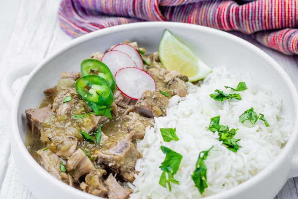 A bowl of white rice and green chili pork stew garnished with cilantro, radish slices, jalapeño, and a lime wedge, set on a white surface with a colorful cloth in the background.