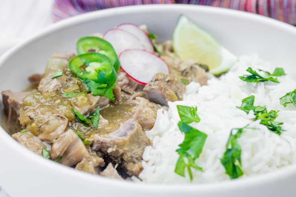 A bowl containing white rice topped with chopped cilantro, and green chili pork stew garnished with sliced radishes, jalapeños, cilantro, and a lime wedge.