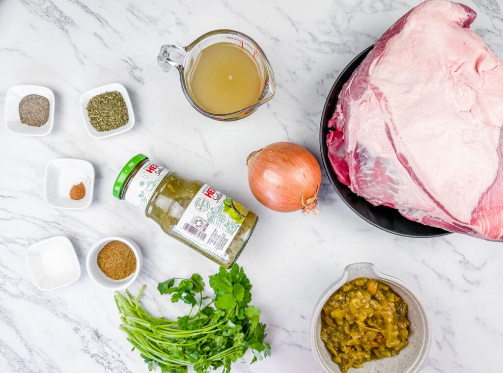 Raw brisket, an onion, beef broth, jar of salsa verde, chopped green chiles, cilantro, and various spices arranged on a white marble surface.