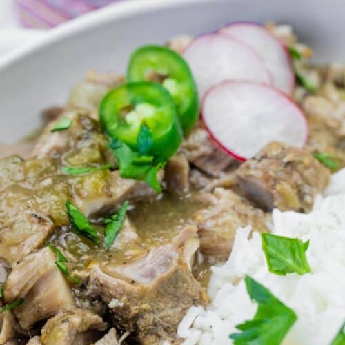 A close-up of a bowl with stewed meat in green sauce, white rice, and garnished with sliced jalapeños, radishes, and fresh herbs.