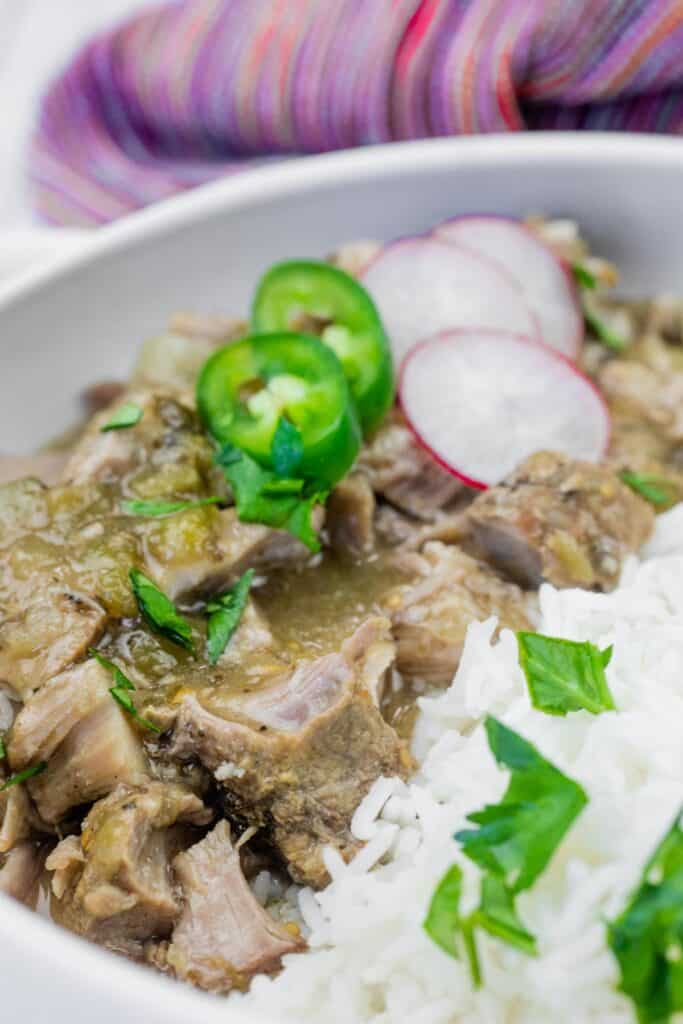 A close-up of a bowl with stewed meat in green sauce, white rice, and garnished with sliced jalapeños, radishes, and fresh herbs.