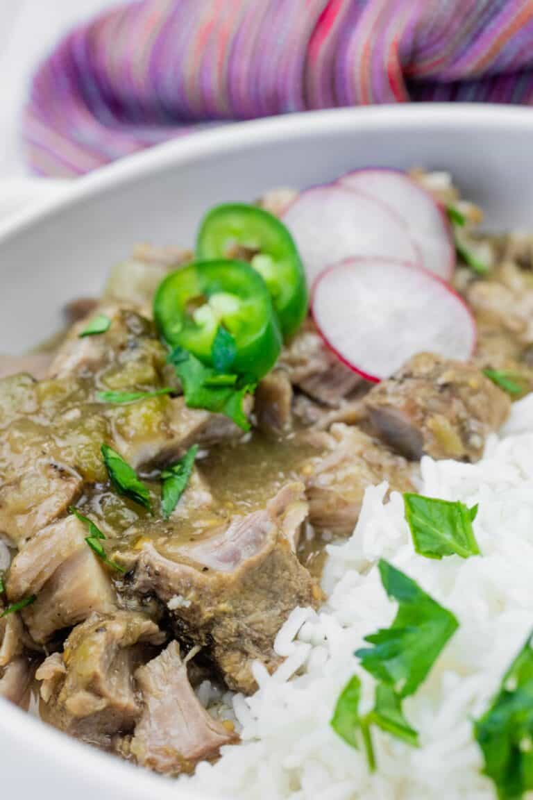 A close-up of a bowl with stewed meat in green sauce, white rice, and garnished with sliced jalapeños, radishes, and fresh herbs.