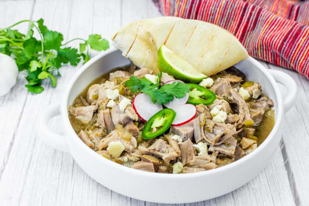 A bowl of pork stew topped with radish, jalapeño, cilantro, lime, and crumbled cheese, served with a slice of grilled bread; cilantro garnish and a cloth napkin in the background.