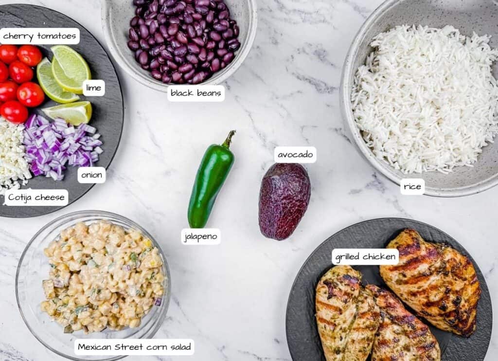 Overhead shot of street corn chicken rice bowl ingredients, including chicken, rice, beans, corn salad, avocado, and vegetables, arranged on a light surface with labels for each ingredient.