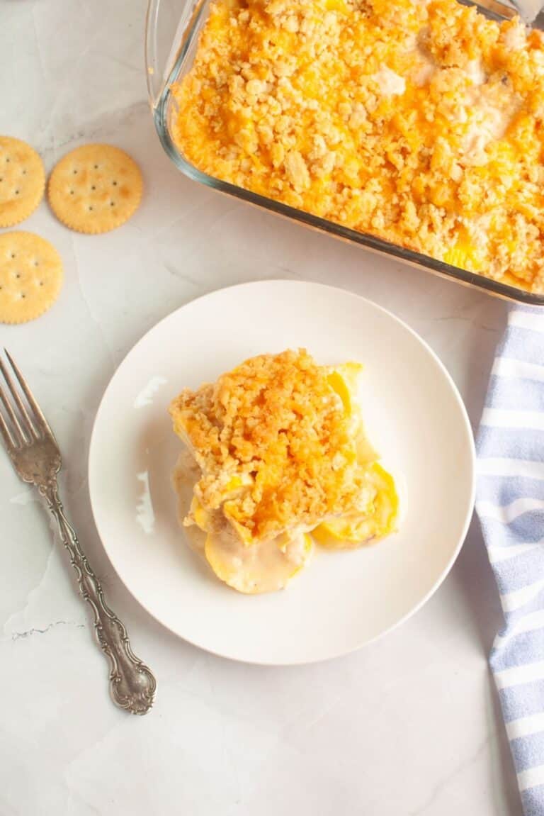 A serving of cheesy squash casserole with a crumb topping on a white plate, next to a fork, crackers, and a casserole dish on a light surface.