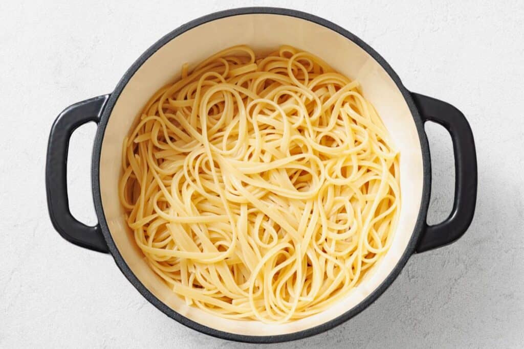 A large black pot filled with cooked plain linguine pasta sits on a light-colored surface.