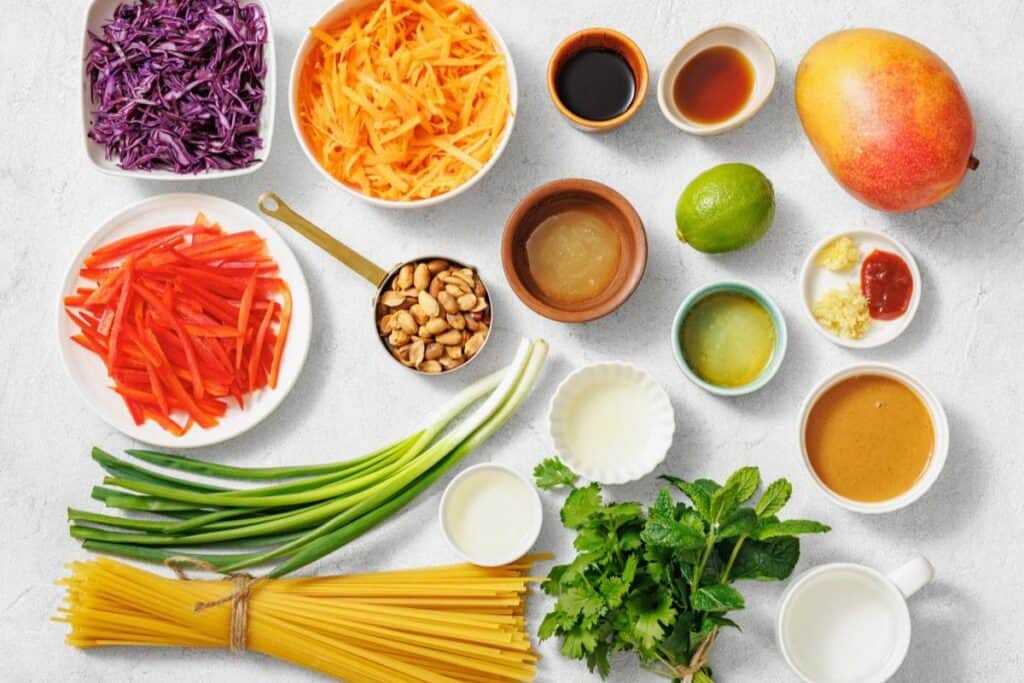 Ingredients arranged on a counter including uncooked pasta, shredded vegetables, herbs, lime, sauces, peanuts, a mango, and various seasonings in small bowls.