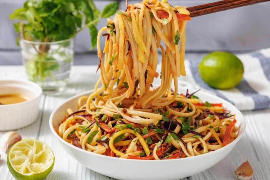 A bowl of vegetable noodle salad is lifted with chopsticks, surrounded by fresh lime, herbs, and a small dish of sauce on a light table.