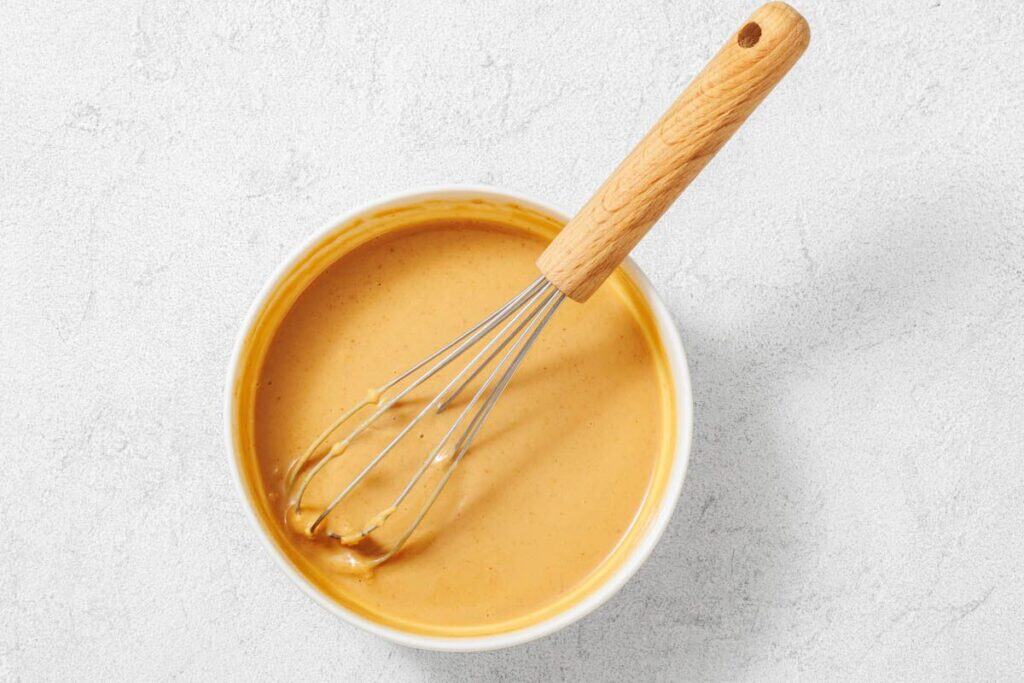 A metal whisk with a wooden handle rests in a bowl of creamy peanut butter mixture on a light-colored surface.