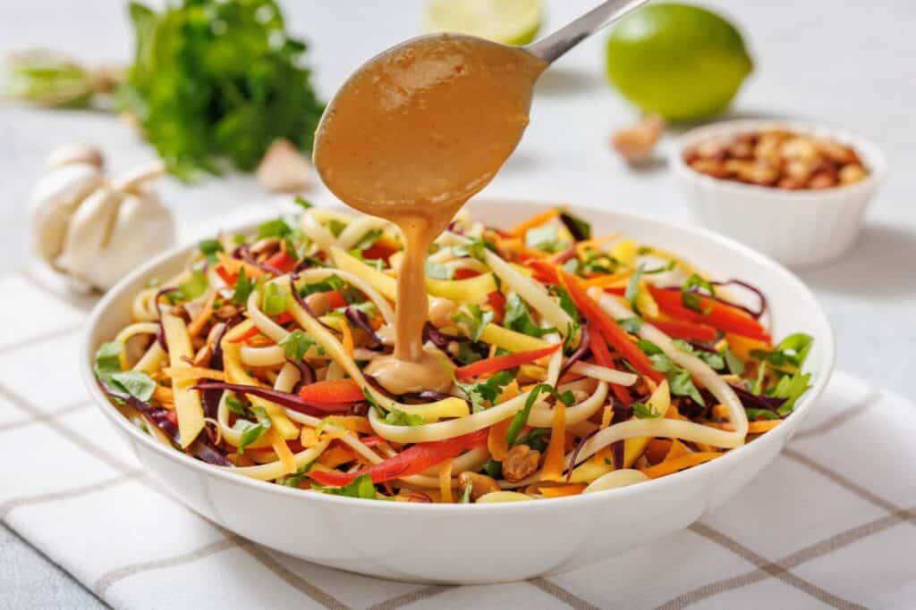 A hand drizzles a creamy dressing over a bowl of colorful vegetable noodle salad with herbs, nuts, and lime in the background.