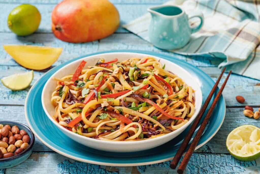 A bowl of noodle salad with julienned vegetables, garnished with chopped peanuts and herbs, placed on a blue plate with chopsticks, surrounded by mango, lime, and a small jug.