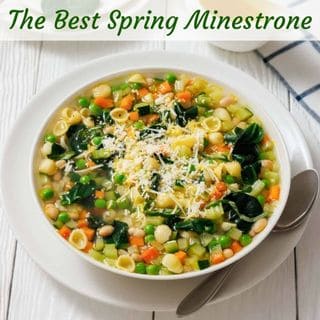 A bowl of spring minestrone soup with pasta, beans, spinach, peas, carrots, and grated cheese, served with a spoon on a white table.