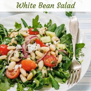 A plate of white bean salad with arugula, cherry tomatoes, red onion, cucumber, capers, and crumbled cheese, served with a fork on the side.