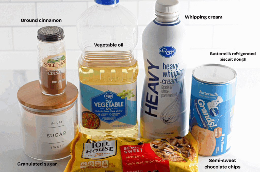 Assorted baking ingredients on a counter: ground cinnamon, granulated sugar, vegetable oil, heavy whipping cream, refrigerated biscuit dough, and semi-sweet chocolate chips.