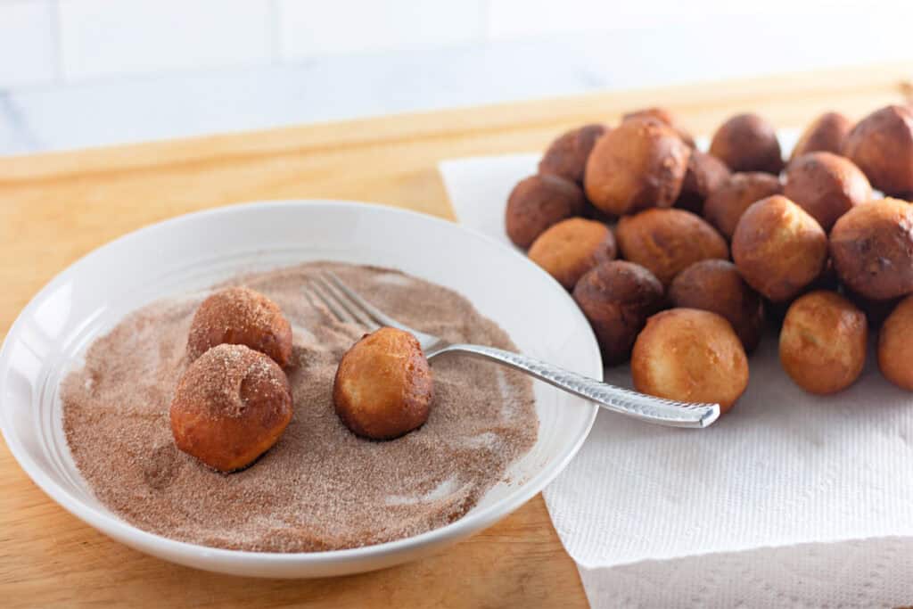 A plate with small fried dough balls being rolled in cinnamon sugar with a fork, next to a pile of plain dough balls on a paper towel.