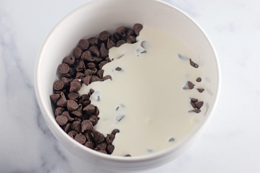 A white bowl containing chocolate chips on one side and cream partially covering them, placed on a light surface.