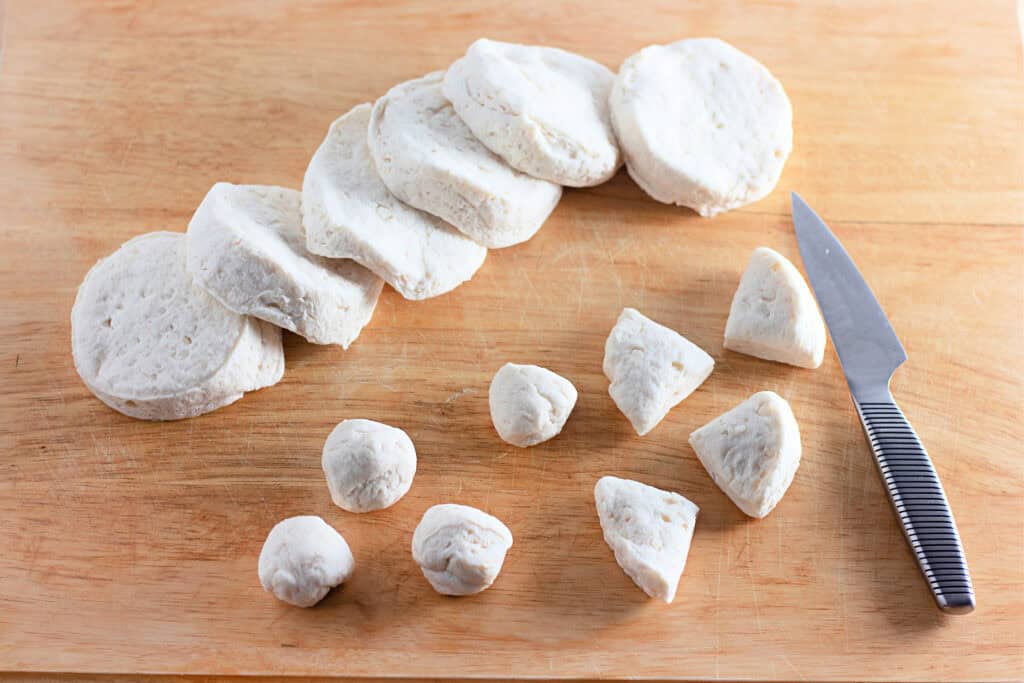 Sliced biscuit dough rounds and dough balls are arranged on a wooden cutting board next to a knife.