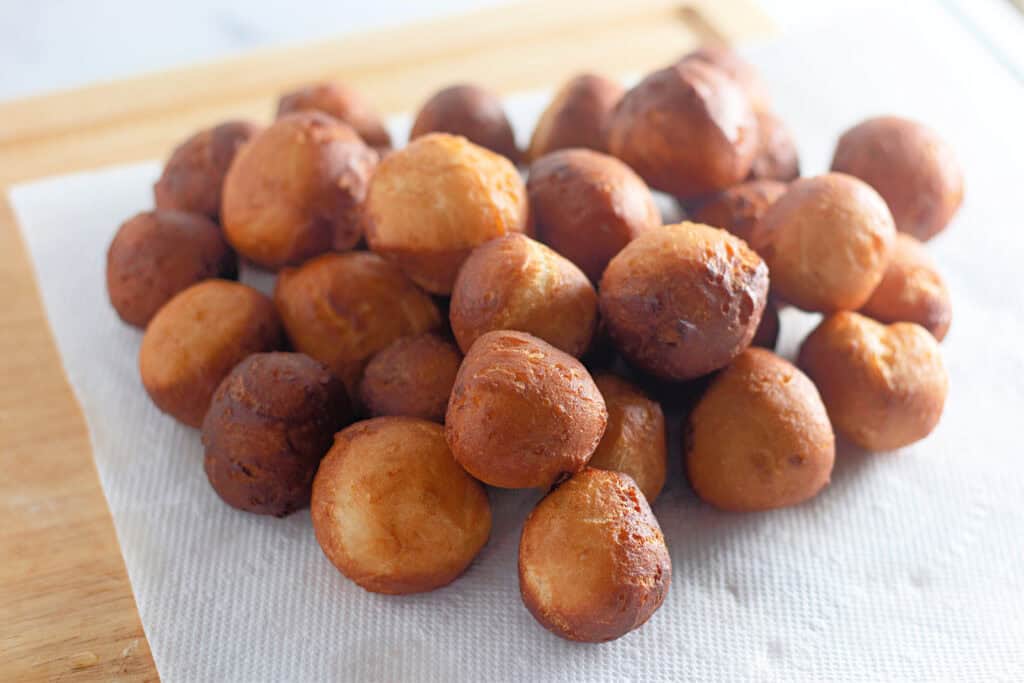 A pile of round, golden-brown fried dough balls resting on a paper towel atop a wooden surface.