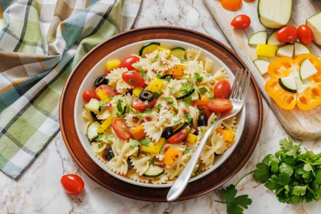 A plate of farfalle pasta salad with cherry tomatoes, zucchini, yellow bell pepper, black olives, and parsley, on a brown-rimmed plate with a fork, beside fresh vegetables and a checkered cloth.