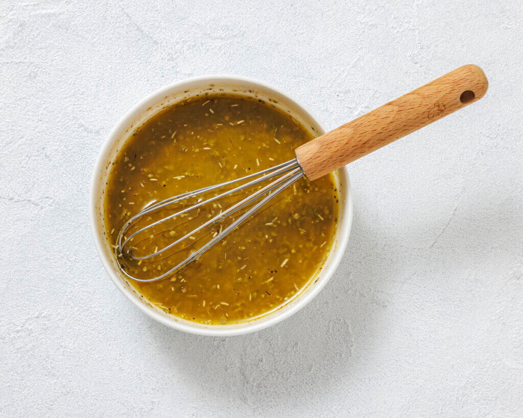 A bowl of yellow-green vinaigrette dressing with herbs and a whisk resting inside, placed on a light textured surface.