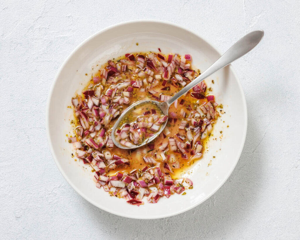 A white bowl containing a mixture of chopped red onions, herbs, and liquid dressing, with a metal spoon resting inside.
