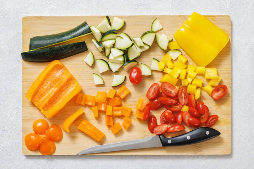 A cutting board with chopped yellow bell pepper, orange bell pepper, zucchini, and halved cherry tomatoes next to a kitchen knife.