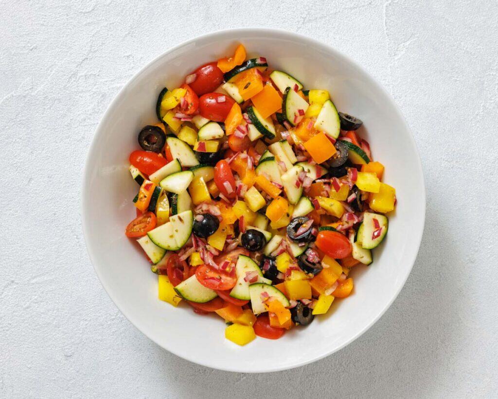 A white bowl filled with a colorful vegetable salad, including cherry tomatoes, yellow bell peppers, zucchini, red onions, and black olives, on a light gray surface.