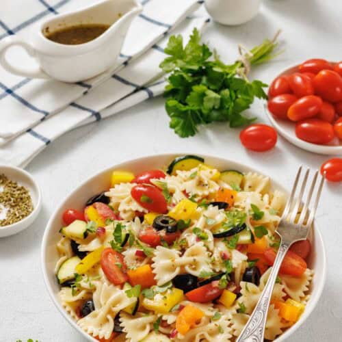 A bowl of pasta salad with bowtie pasta, cherry tomatoes, black olives, yellow bell peppers, and cucumber, with a fork and fresh ingredients around it on a white surface.
