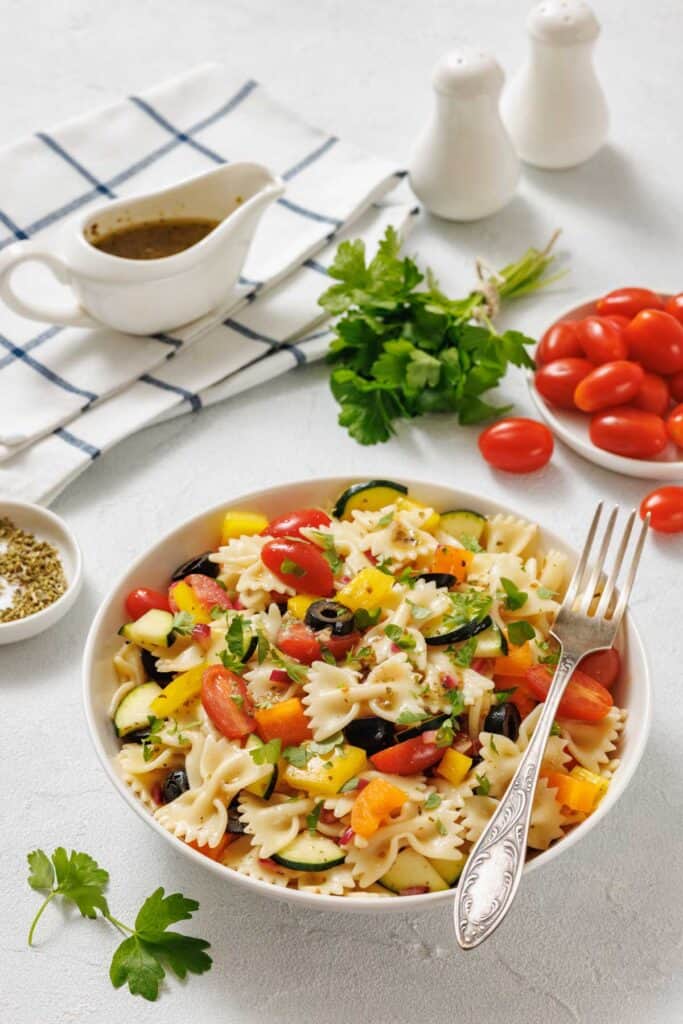 A bowl of pasta salad with bowtie pasta, cherry tomatoes, black olives, yellow bell peppers, and cucumber, with a fork and fresh ingredients around it on a white surface.