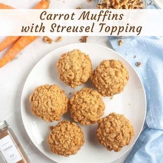 A white plate with six carrot muffins topped with streusel sits on a table next to carrots, walnuts, a spice jar, and a light blue cloth. Text above reads: "Carrot Muffins with Streusel Topping.