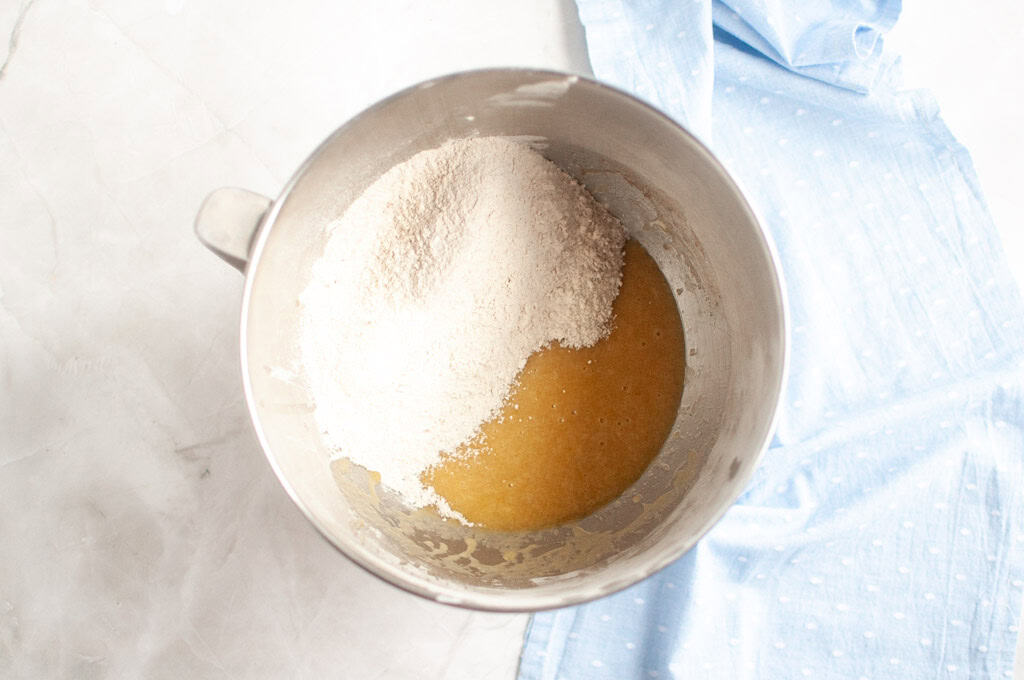 Overhead shot of whole wheat flour being added to wet ingredients in a metal mixing bowl.