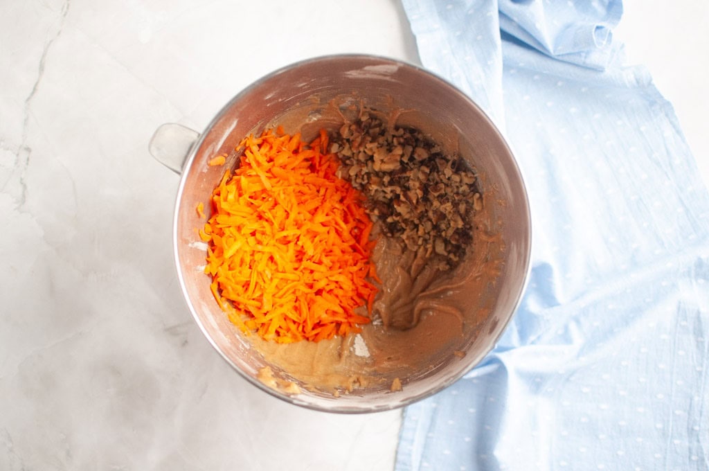 Overhead shot of carrot muffin batter in a metal mixing bowl with shredded carrots and chopped walnuts being added.