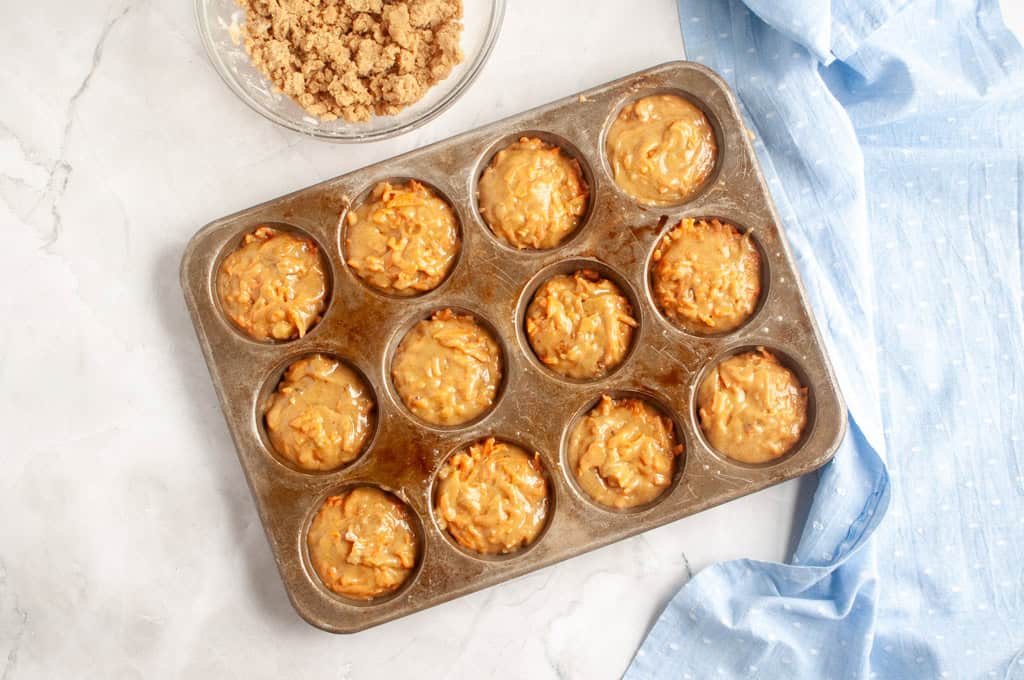 Overhead shot of carrot muffin batter filled in a muffin tin, with streusel topping nearby.
