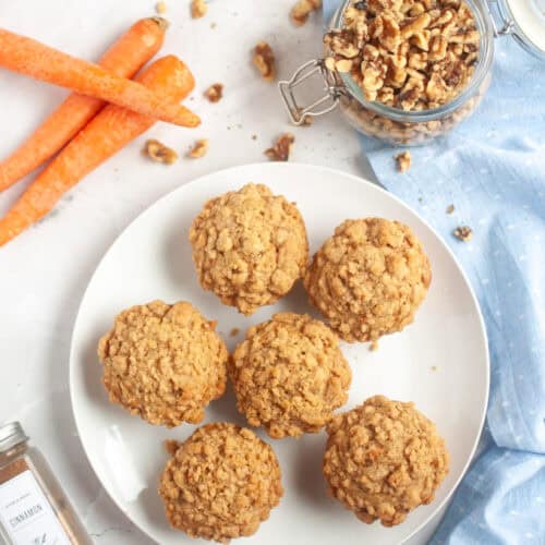 Six golden-brown Carrot muffins arranged on a white plate, with carrots and walnuts nearby.