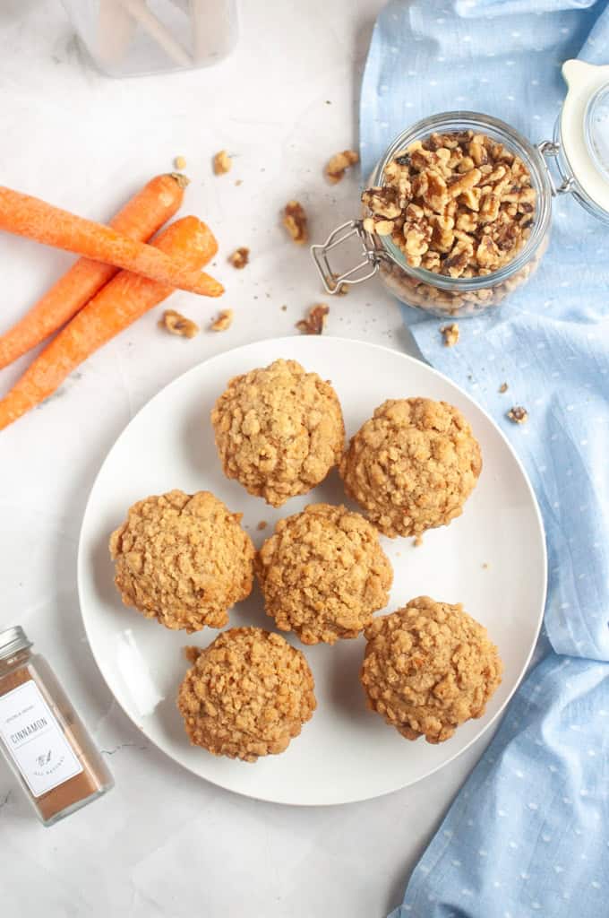 Six golden-brown Carrot muffins arranged on a white plate, with carrots and walnuts nearby.