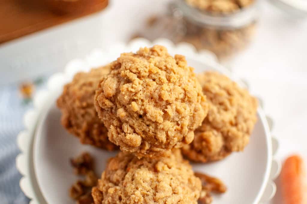 Six carrot muffins with streusel topping on a white plate, surrounded by carrots, cinnamon, and streusel.