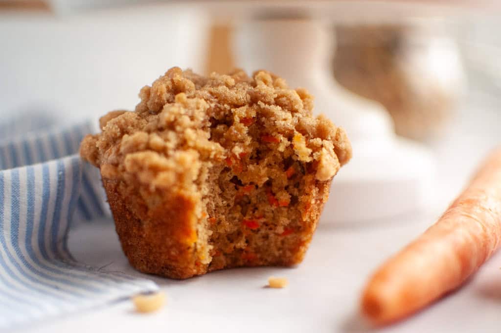 A close-up of a carrot cake muffin with a streusel topping, with a bite missing, next to a carrot.