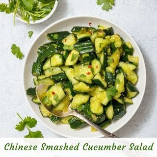 A bowl of chopped cucumber salad with red pepper flakes and cilantro, accompanied by a silver spoon. A small bowl of fresh cilantro is placed next to the salad.