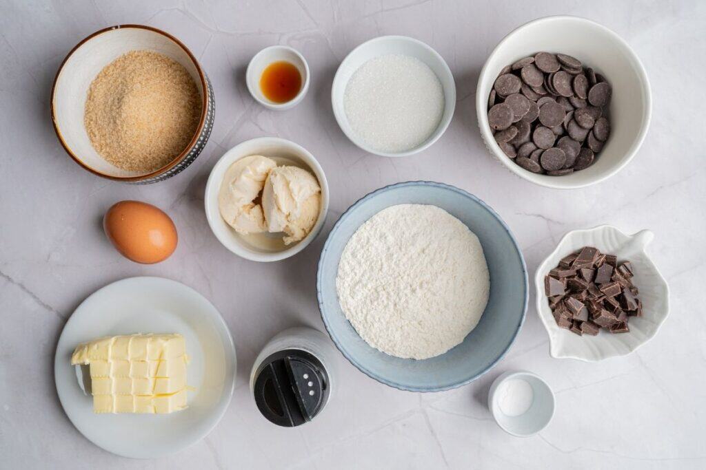 Overhead shot of labeled ingredients for a chocolate chip cookie, including flour, sugar, brown sugar, butter, egg, vanilla, baking soda, salt, and chocolate chips.