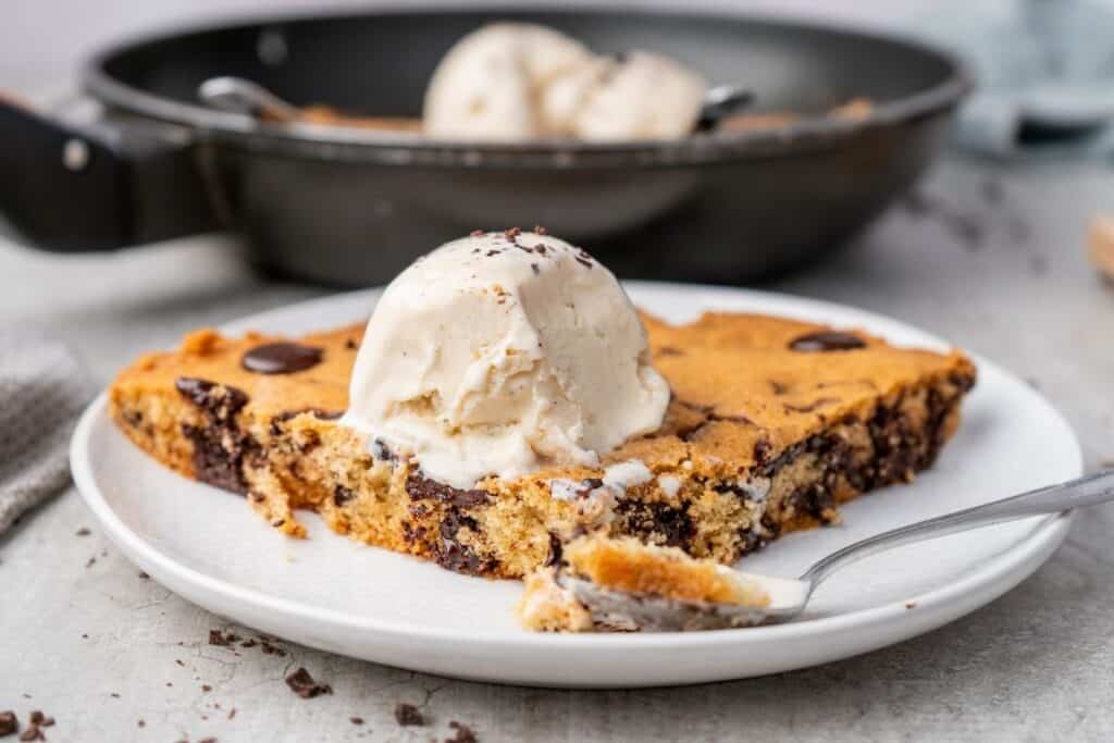 A warm chocolate chip skillet cookie in a black skillet, topped with scoops of vanilla ice cream.