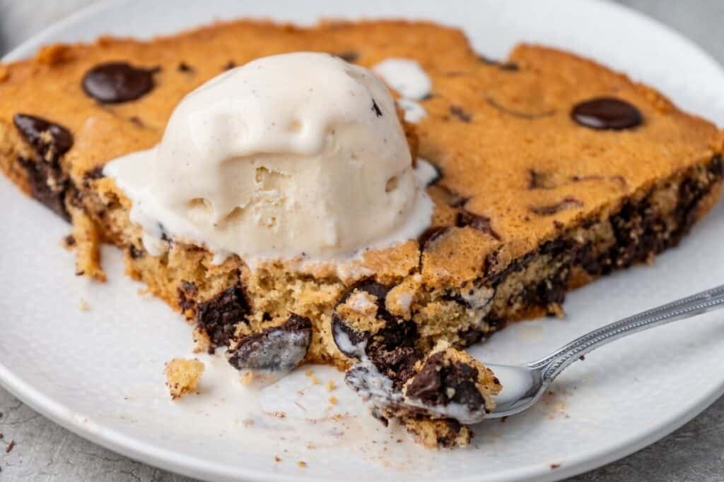A close-up of a slice of warm chocolate chip skillet cookie topped with melting vanilla ice cream on a white plate, with a bite on a spoon.