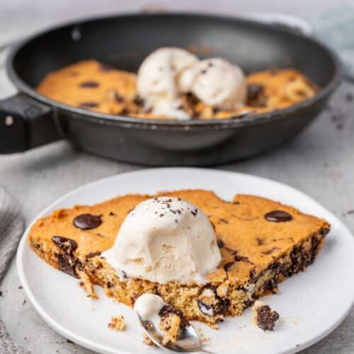A slice of warm chocolate chip skillet cookie topped with melting vanilla ice cream on a white plate.