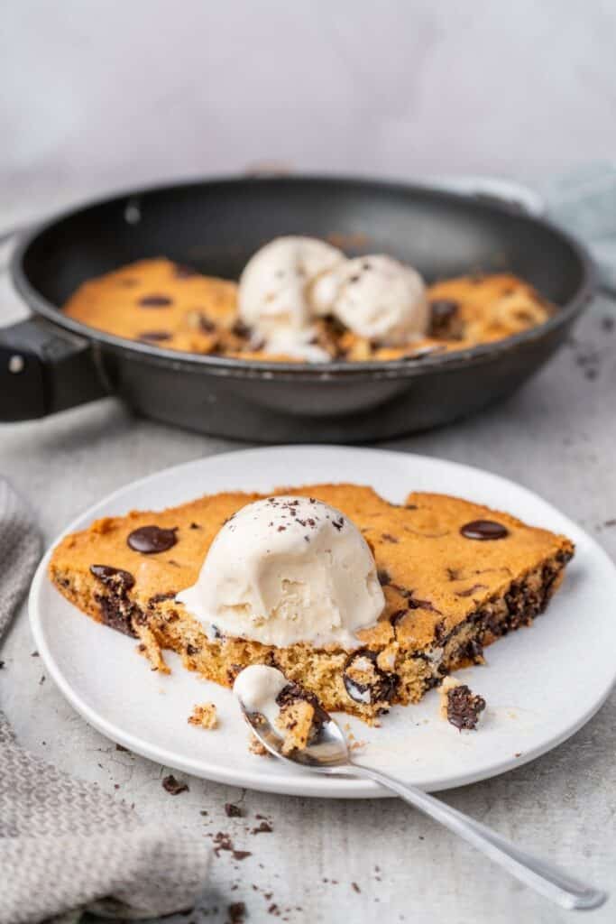 A slice of warm chocolate chip skillet cookie topped with melting vanilla ice cream on a white plate.