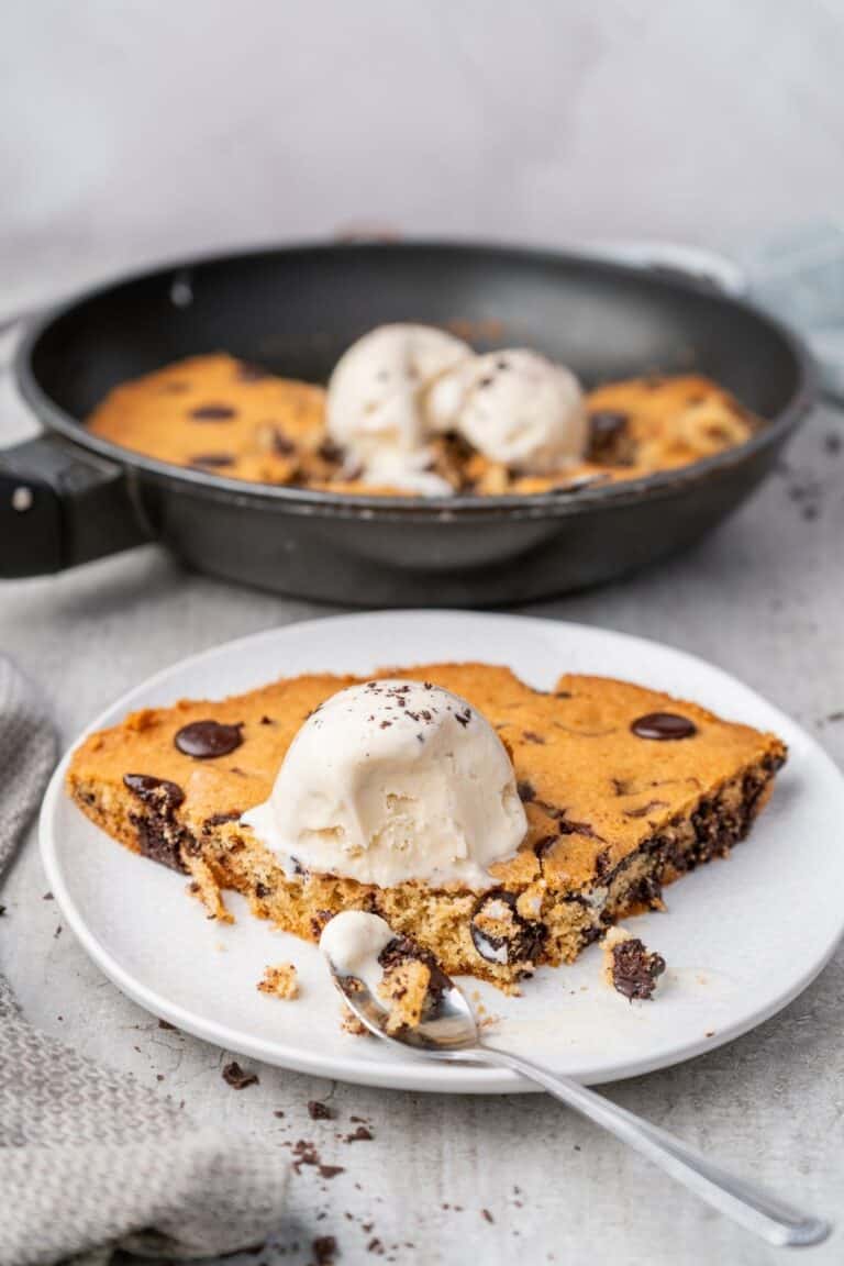 A slice of warm chocolate chip skillet cookie topped with melting vanilla ice cream on a white plate.