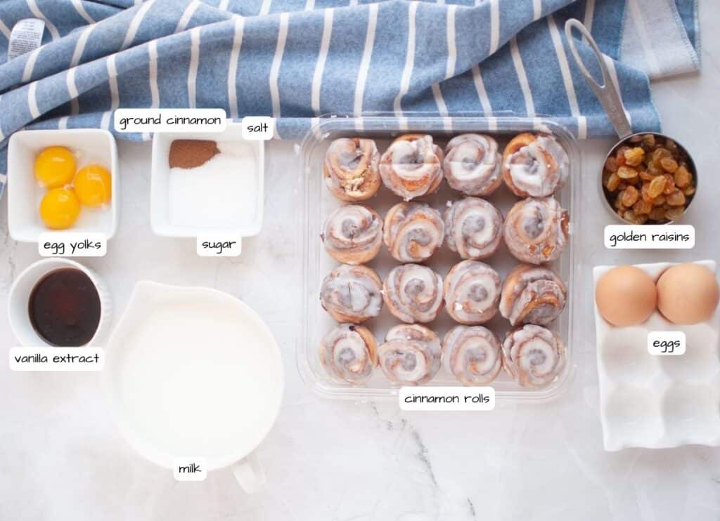 Overhead shot of labeled ingredients for cinnamon roll casserole including cinnamon rolls, eggs, egg yolks, milk, vanilla extract, ground cinnamon, sugar, salt, and golden raisins.